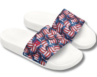 May include: White slide sandals featuring a baseball-themed design. The straps display a pattern of baseballs with red and blue accents, resembling the American flag. The sandals are designed for casual wear.