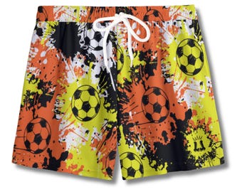 May include: Soccer-themed swim trunks with a vibrant design. The shorts feature a repeating pattern of soccer balls and paint splatters in yellow, orange, black, and white. They have a white drawstring and elastic waistband for a comfortable fit.
