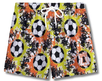 May include: Soccer-themed swim trunks featuring a repeating pattern of black and white soccer balls on a yellow, orange, and black splatter background. The shorts have a white drawstring and are likely made of a lightweight, quick-drying material.