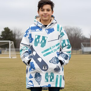 May include: White hooded blanket with a blue and teal winter sports pattern. The blanket features images of ice skates, hockey sticks, gloves, and curling stones. The blanket has a fleece lining and a large front pocket.