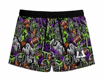 May include: Black athletic shorts featuring a vibrant all-over print of a graveyard scene with tombstones, skulls, ghosts, footballs, and goalposts. The design incorporates neon green, purple, and orange accents with black drawstrings.