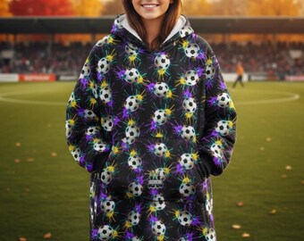 May include: A black hooded blanket with a soccer ball pattern and colorful splatter designs. The blanket has a white interior and a large front pocket. The person is standing on a green field.