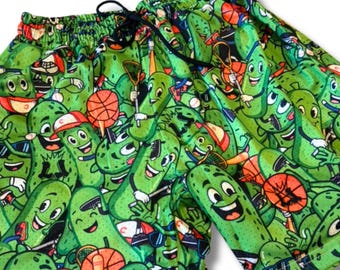 May include: Green shorts with a repeating pattern of cartoon pickles wearing sunglasses, hats, and playing sports. The shorts have a drawstring waistband and are made of a lightweight fabric.