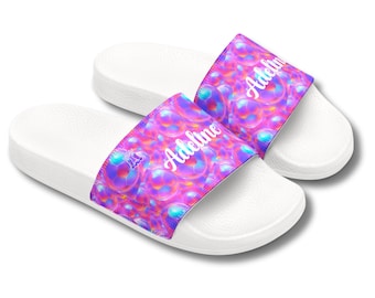 May include: White slide sandals with a colorful bubble design on the straps. The straps feature the name "Adeline" in a cursive font. The sandals are designed for casual wear and are set against a white background.