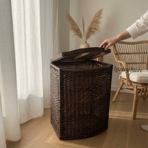 Puede incluir: Una cesta de almacenamiento tejida de color marrón oscuro con una tapa con bisagras. La cesta es rectangular con esquinas redondeadas y se apoya en un suelo de madera clara. La tapa está parcialmente abierta, revelando el interior. La cesta está junto a una ventana con cortinas blancas.