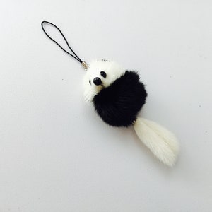 May include: A black and white fur pom-pom keychain with a black cord. The pom-pom has a white face with black eyes and a black nose.