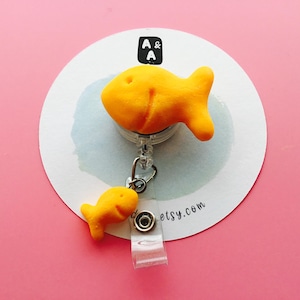 May include: A novelty badge reel featuring two bright yellow goldfish crackers. The larger fish is attached to the reel, and a smaller fish dangles below. The reel is mounted on a white circular card with the text "A & A" and "etsy.com".