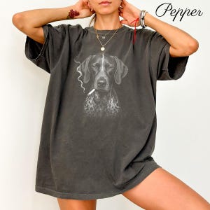 Può includere: Una t-shirt oversize grigio antracite con una stampa grafica di un bracco tedesco a pelo corto con una sigaretta in bocca e una scia di fumo. La parola Pepper è stampata nell'angolo in alto a destra con un carattere corsivo nero.