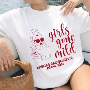 May include: A white short-sleeved t-shirt featuring a red graphic of a person wearing a towel on their head and sunglasses, making a peace sign. The text reads girls gone mild and AMELIA'S BACHELORETTE MIAMI, 2025 in a red script and block font.
