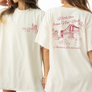 May include: Cream-coloured t-shirt with red graphics. The front displays "The Bachelorette Club" and a Statue of Liberty illustration. The back features a New York theme with the text "With love from New York" and "Amelia's Bachelorette", plus a bridge and taxi.