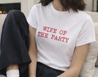 May include: White t-shirt with the red text "WIFE OF THE PARTY." The shirt has short sleeves and a crew neck. The model is wearing navy blue pants and white socks.