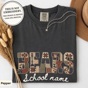 May include: A charcoal gray t-shirt featuring the word BEARS in a patchwork quilt pattern with various floral and geometric prints, and the text School name below. The shirt includes a tag reading Comfort Colors and a silver star necklace.