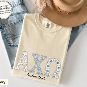 Può includere: Una t-shirt color avorio con le lettere greche Alpha Chi Omega in una stampa floreale blu con un contorno beige. La maglietta include il testo "Custom text" sotto le lettere e un'etichetta circolare che recita "THIS IS NOT EMBROIDERY. THIS DESIGN IS PRINTED USING DTG PRINTING."