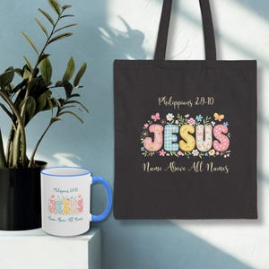May include: A black tote bag and a white mug with a blue handle and rim. Both items feature the word "JESUS" in colorful, floral-style lettering. The tote bag has the text "Philippians 2:9-10" and "Name Above All Names".