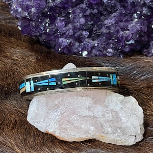 May include: Silver-toned bracelet with a black inlay and colorful geometric designs. The bracelet is resting on a light pink crystal, with a large purple amethyst crystal in the background. The bracelet's design includes blue, turquoise, and orange accents.