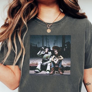 May include: A charcoal gray short-sleeve t-shirt featuring a graphic print of the characters Buzz Lightyear and Woody sitting together on a curb at night. The graphic shows the two figures in a dark outdoor setting with utility poles in the background.