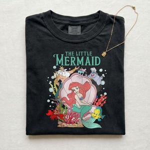Op de afbeelding: Zwart T-shirt met een kleurrijke afbeelding van Ariel uit De Kleine Zeemeermin, met de tekst "THE LITTLE MERMAID" in teal. Het ontwerp bevat personages als Botje, Sebastiaan en Koning Triton. Een gouden ketting met een kleine hanger ligt op het shirt.