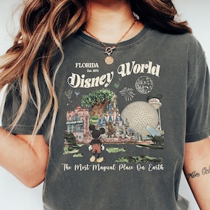 Puede incluir: Camiseta gris oscuro con un dise&ntilde;o vintage de Disney World. El dise&ntilde;o incluye el nombre del parque, la fecha de fundaci&oacute;n y lugares emblem&aacute;ticos como el castillo de Cenicienta y Spaceship Earth, con Mickey Mouse. El texto dice "The Most Magical Place On Earth."