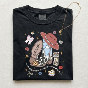 Może przedstawiać: Czarny t-shirt z nadrukiem graficznym przedstawiającym kowbojski but w łaty, czerwony kowbojski kapelusz oraz klocki z napisem BTS. Na bucie widnieje imię Taylor i liczba 13, otoczone gwiazdkami, kwiatami i kokardkami.