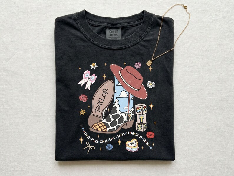 Może przedstawiać: Czarny t-shirt z nadrukiem graficznym przedstawiającym kowbojski but w łaty, czerwony kowbojski kapelusz oraz klocki z napisem BTS. Na bucie widnieje imię Taylor i liczba 13, otoczone gwiazdkami, kwiatami i kokardkami.