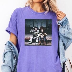 May include: A purple short-sleeved t-shirt featuring a square graphic print of two animated characters sitting on a curb at night. One character is a space ranger in a white and green suit, and the other is a cowboy in a brown hat and vest.