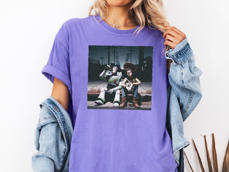 May include: A purple short-sleeved t-shirt featuring a square graphic print of two animated characters sitting on a curb at night. One character is a space ranger in a white and green suit, and the other is a cowboy in a brown hat and vest.