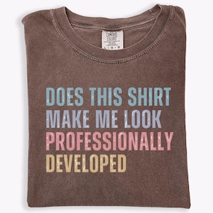 May include: A brown crew neck t-shirt with a distressed graphic print that reads "DOES THIS SHIRT MAKE ME LOOK PROFESSIONALLY DEVELOPED" in stacked lines of light blue, light purple, pink, and yellow text. The shirt features a Comfort Colors label.
