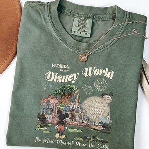 Puede incluir: Una camiseta verde oliva con un gr&aacute;fico de Disney World en Florida. El dise&ntilde;o incluye a Mickey Mouse, un castillo y el texto "Disney World" y "The Most Magical Place On Earth". Tambi&eacute;n se ven un collar dorado y un sombrero marr&oacute;n.