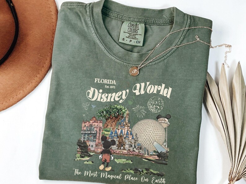 Puede incluir: Una camiseta verde oliva con un gr&aacute;fico de Disney World en Florida. El dise&ntilde;o incluye a Mickey Mouse, un castillo y el texto "Disney World" y "The Most Magical Place On Earth". Tambi&eacute;n se ven un collar dorado y un sombrero marr&oacute;n.