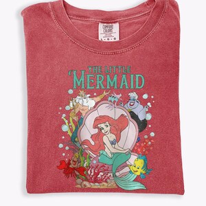 Op de afbeelding: Rood Comfort Colors T-shirt met een afbeelding van Ariel uit De Kleine Zeemeermin. Het ontwerp bevat Ariel, Botje, Sebastiaan, Koning Triton en Ursula. De woorden "THE LITTLE MERMAID" staan in turquoise letters.