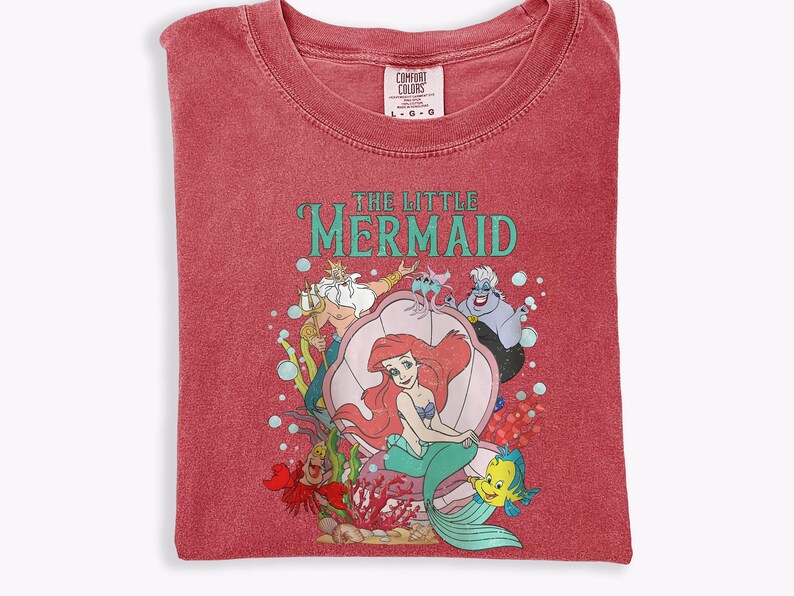 Op de afbeelding: Rood Comfort Colors T-shirt met een afbeelding van Ariel uit De Kleine Zeemeermin. Het ontwerp bevat Ariel, Botje, Sebastiaan, Koning Triton en Ursula. De woorden "THE LITTLE MERMAID" staan in turquoise letters.