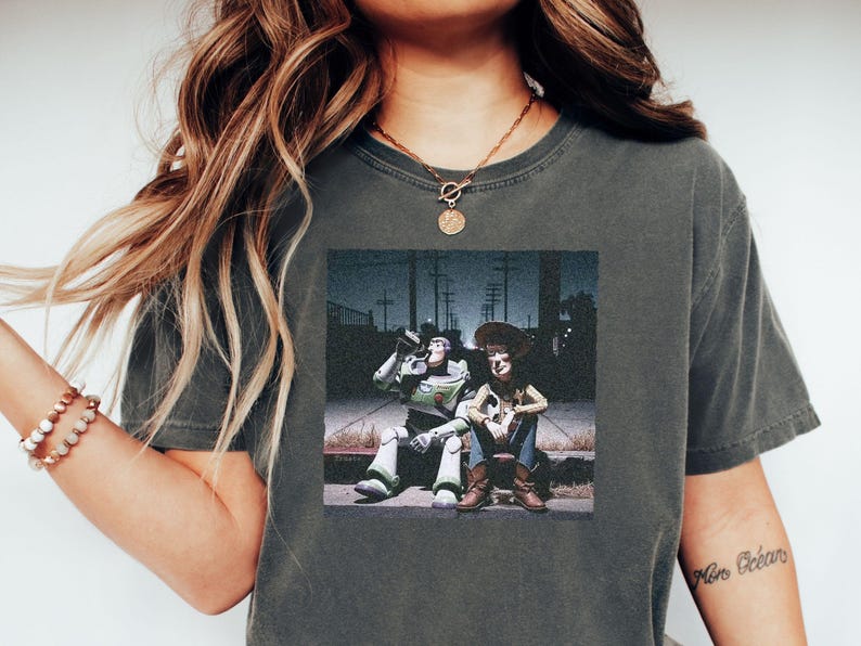 May include: A charcoal gray short-sleeve t-shirt featuring a graphic print of the characters Buzz Lightyear and Woody sitting together on a curb at night. The graphic shows the two figures in a dark outdoor setting with utility poles in the background.
