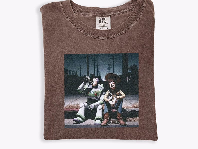 May include: A brown short-sleeve t-shirt featuring a graphic print of Buzz Lightyear and Woody sitting on a curb at night. The shirt has a crew neckline and a Comfort Colors brand label on the inside collar, indicating a size large.