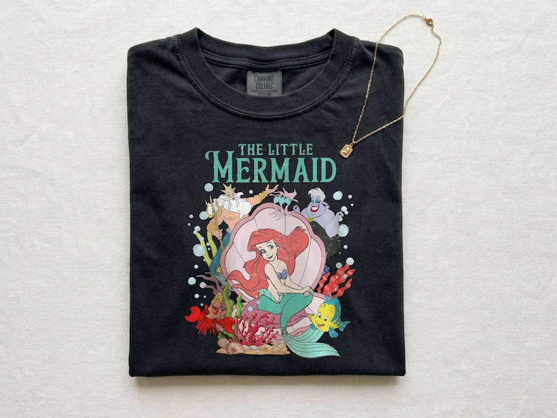 Op de afbeelding: Zwart T-shirt met een kleurrijke afbeelding van Ariel uit De Kleine Zeemeermin, met de tekst "THE LITTLE MERMAID" in teal. Het ontwerp bevat personages als Botje, Sebastiaan en Koning Triton. Een gouden ketting met een kleine hanger ligt op het shirt.