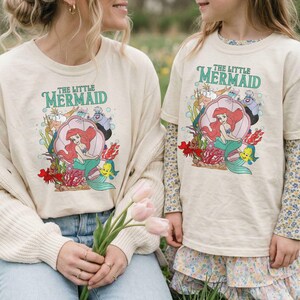 Op de afbeelding: Twee cr&egrave;mekleurige T-shirts met de tekst "The Little Mermaid" in het groen. De shirts tonen een kleurrijke afbeelding van Ariel, Botje en Sebastiaan in een schelp. E&eacute;n shirt heeft lange mouwen, de andere korte mouwen.