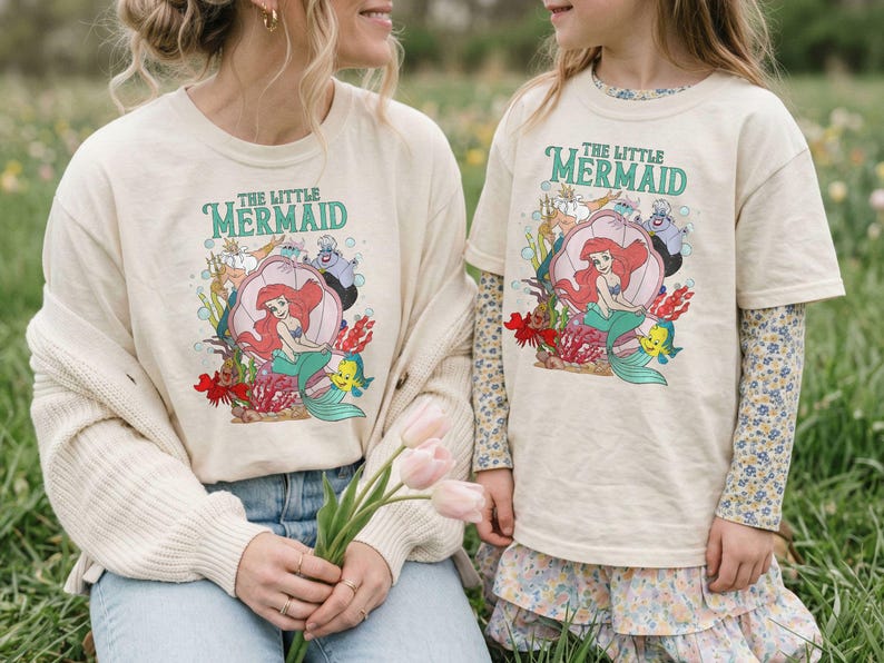 Op de afbeelding: Twee cr&egrave;mekleurige T-shirts met de tekst "The Little Mermaid" in het groen. De shirts tonen een kleurrijke afbeelding van Ariel, Botje en Sebastiaan in een schelp. E&eacute;n shirt heeft lange mouwen, de andere korte mouwen.