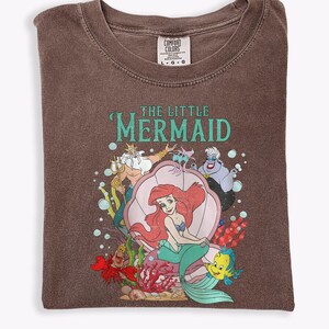 Op de afbeelding: Bruin T-shirt met een afbeelding van Ariel uit De Kleine Zeemeermin. Het ontwerp bevat Ariel in een zeeschelp, met Koning Triton, Botje, Sebastiaan en Ursula. De tekst "THE LITTLE MERMAID" is turquoise.