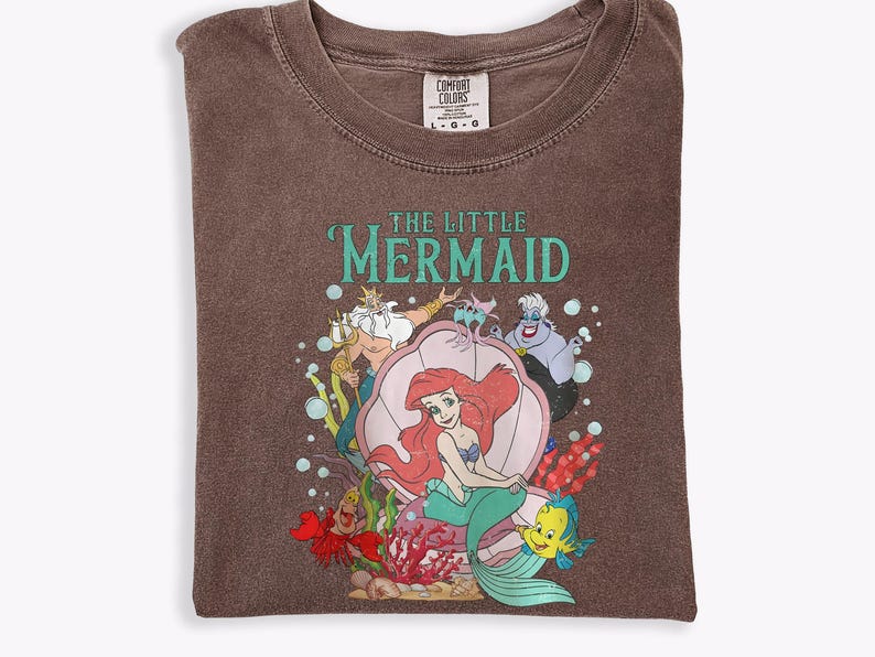 Op de afbeelding: Bruin T-shirt met een afbeelding van Ariel uit De Kleine Zeemeermin. Het ontwerp bevat Ariel in een zeeschelp, met Koning Triton, Botje, Sebastiaan en Ursula. De tekst "THE LITTLE MERMAID" is turquoise.