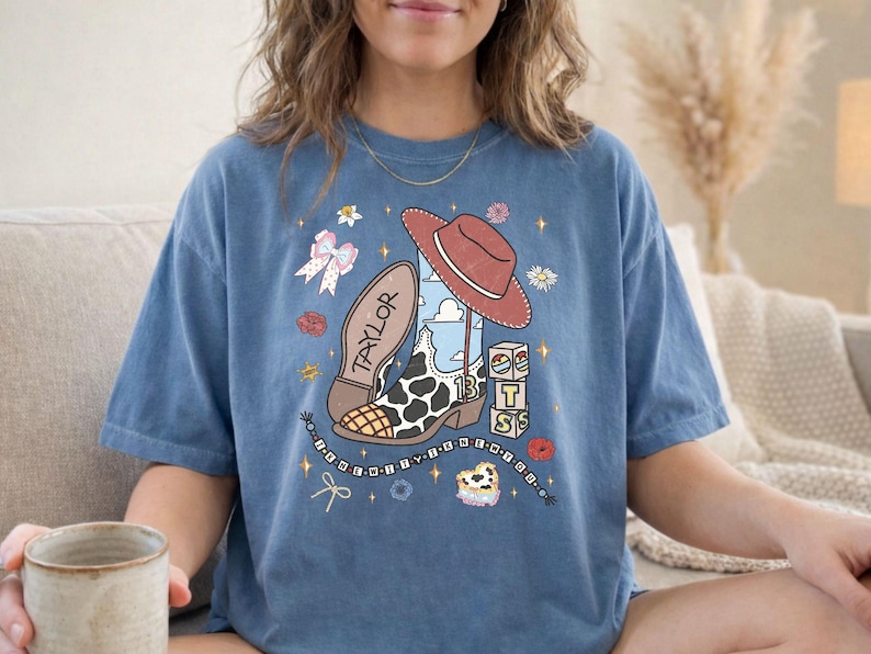 Może przedstawiać: Niebieski t-shirt z kr&oacute;tkim rękawem z nadrukiem graficznym przedstawiającym kowbojski but w łaty, czerwony kapelusz, klocki z literami tworzącymi napis BTS oraz imię Taylor, otoczone małymi gwiazdkami, kwiatami, kokardą i sznurkiem koralik&oacute;w.