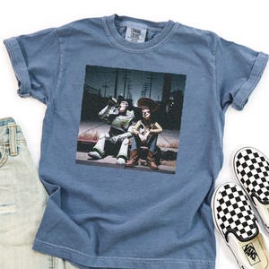 May include: A blue short-sleeve t-shirt featuring a graphic print of Buzz Lightyear and Woody sitting on a curb at night. The shirt is displayed alongside a pair of light-wash denim jeans and black and white checkered slip-on sneakers.