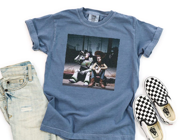 May include: A blue short-sleeve t-shirt featuring a graphic print of Buzz Lightyear and Woody sitting on a curb at night. The shirt is displayed alongside a pair of light-wash denim jeans and black and white checkered slip-on sneakers.