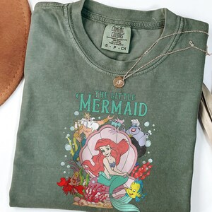 Op de afbeelding: Olijfgroen T-shirt met een afbeelding van Ariel uit De Kleine Zeemeermin, met de tekst "THE LITTLE MERMAID". Een gouden ketting met een hanger ligt op het shirt. Een bruine hoed en een gedroogde plant staan op de achtergrond.