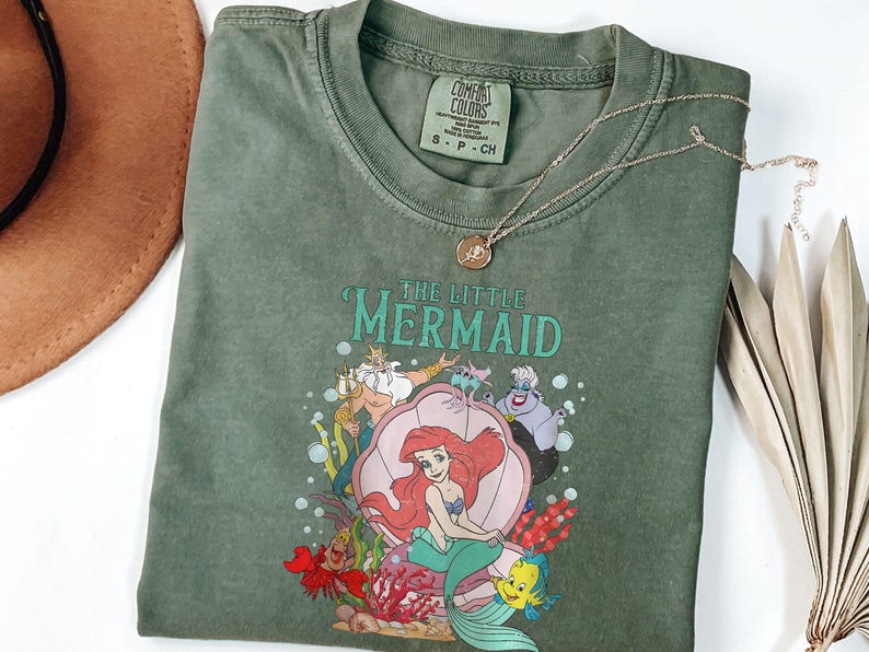 Op de afbeelding: Olijfgroen T-shirt met een afbeelding van Ariel uit De Kleine Zeemeermin, met de tekst "THE LITTLE MERMAID". Een gouden ketting met een hanger ligt op het shirt. Een bruine hoed en een gedroogde plant staan op de achtergrond.