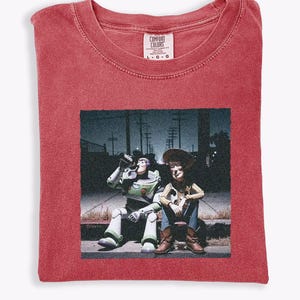 May include: A red short-sleeve t-shirt with a crew neckline and a Comfort Colors brand label at the collar. The center features a graphic print of Buzz Lightyear and Woody sitting on a curb at night, with a dark urban background and streetlights.