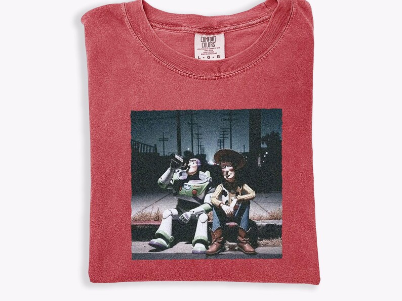 May include: A red short-sleeve t-shirt with a crew neckline and a Comfort Colors brand label at the collar. The center features a graphic print of Buzz Lightyear and Woody sitting on a curb at night, with a dark urban background and streetlights.