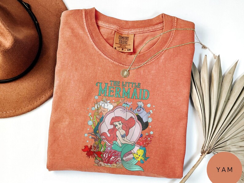 Op de afbeelding: Een koraalkleurig T-shirt met de tekst "THE LITTLE MERMAID" en een afbeelding van Ariel, Botje, Sebastiaan en andere personages. Het shirt heeft een ronde hals en een klein label linksboven. Een gouden ketting en een hoed liggen naast het shirt.
