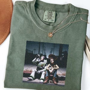 May include: An olive green crew neck t-shirt featuring a graphic print of two animated characters sitting on a curb at night. The shirt has a Comfort Colors brand label at the inner neckline. A thin gold-toned chain necklace rests on the fabric.