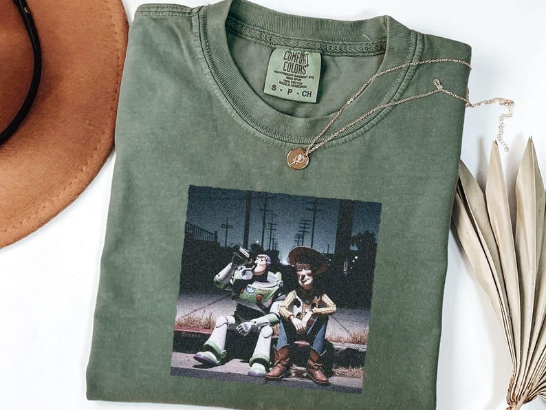 May include: An olive green crew neck t-shirt featuring a graphic print of two animated characters sitting on a curb at night. The shirt has a Comfort Colors brand label at the inner neckline. A thin gold-toned chain necklace rests on the fabric.