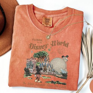 Puede incluir: Una camiseta de color coral con un dise&ntilde;o de Disney World en Florida. El dise&ntilde;o incluye a Mickey Mouse, el &Aacute;rbol de la Vida y Spaceship Earth. El texto dice "Disney World" y "The Most Magical Place on Earth".