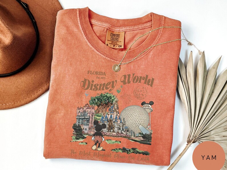 Puede incluir: Una camiseta de color coral con un dise&ntilde;o de Disney World en Florida. El dise&ntilde;o incluye a Mickey Mouse, el &Aacute;rbol de la Vida y Spaceship Earth. El texto dice "Disney World" y "The Most Magical Place on Earth".